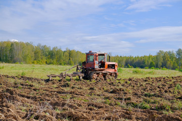 A tracked tractor with a plough plows the ground in autumn against the background of the forest.