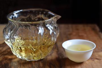 Closeup of a glass small teapot with golden green tea and a small Chinese teacup at a Chinese tea ceremony on a wooden table in front of a dark background