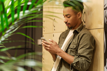 Side view of young casual African American guy thoughtfully using cellphone in locker room alone