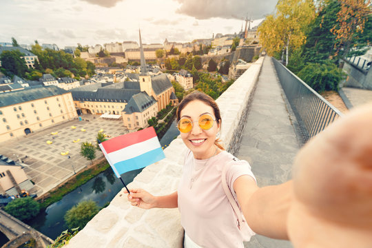 Asian tourist girl takes a selfie with the flag of Luxembourg against the background of the old quarter of the city of Grund and the Church.