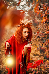 Naklejka premium Woman in long red dress with deer horns in autumn forest. A woman in the autumn forest walks with a lantern and looking for magic herbs. The concept of a fairy tale.