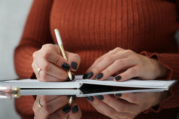 Zbliżenie na dłonie kobiety ubranej w sweterek w jesiennym odcieniu brązu, w trakcie notowania w zeszycie leżącym na biurku ze szklanym blatem
