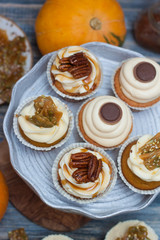 Pumpkin cupcakes with cream cheese frosting decorated with pecan nuts, maple syrup, caramel bites and chocolate toffee candies on wooden rustic background