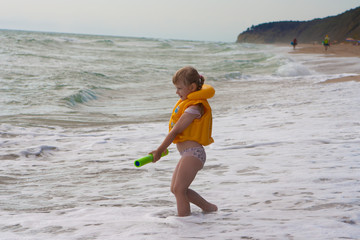 A little girl in a yellow life jacket on the beach plays with a water cannon.