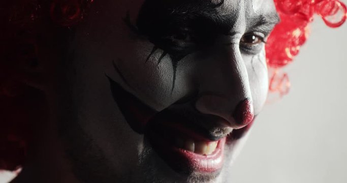Portrait of sad and evil smiling scary jester with wicked frightening humor. Face of angry scary clown turning head and looking at camera at Halloween. Young guy with expressive horror emotions.