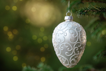 Close-up of white Christmas decoration toy hanging on a spruce branch with bokeh blured lights on background. Copy space.