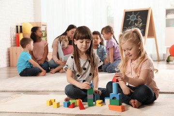 Cute girls playing with building blocks on floor while kindergarten teacher reading book to other...