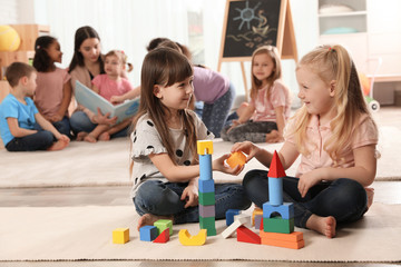 Cute girls playing with building blocks on floor while kindergarten teacher reading book to other...