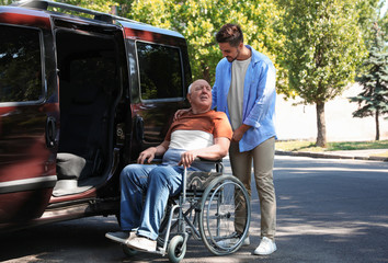 Young man helping patient in wheelchair to get into van outdoors