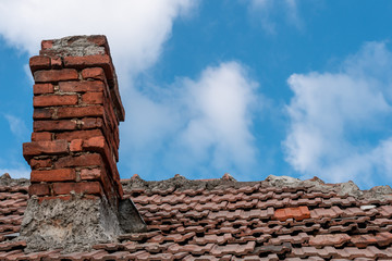 Old red terracotta tile roof with a damaged traditional large brick chimney in need of repair, on a blue sky background. Home winter preparations.