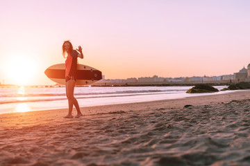 Obraz premium Athletic young girl holds a surfboard and stands at sunset on a sandy beach