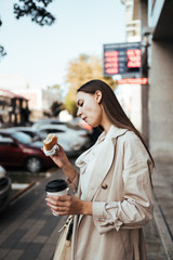 Young business girl in the beige coat eats on the go, holding a roll in her right hand and a paper cup in her left hand, rushing to a meeting