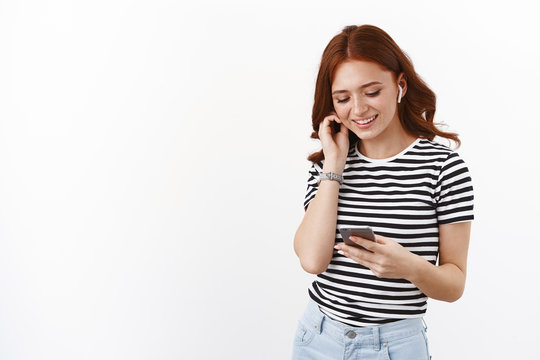Cute redhead teenage girl in striped t-shirt put on wireless earphones, hold smartphone, smiling relaxed and happy mobile phone display, pick song on internet music platform, listen favorite podcast
