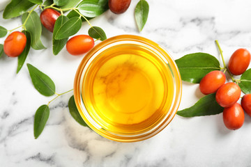Glass bowl with jojoba oil and seeds on white marble table, flat lay