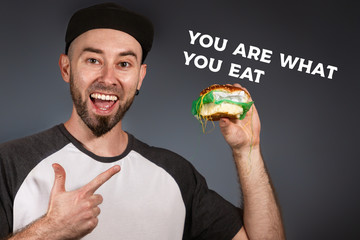 The concept of fast food and ecology. A man in a cap and a beard happily points to a hamburger in...