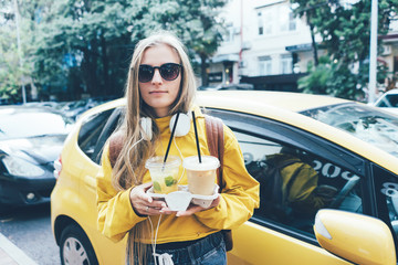 Street portrait of a young stylish woman in a yellow hoody against the background of a yellow car holding refreshments in plastic cups.