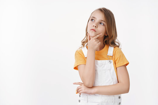 Clever creative pretty young blond little girl look up thoughtful, touch chin making choice, serious gaze top copy space, ponder choice, solving puzzle, stand white background focused, thinking