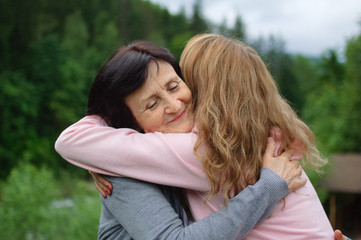 Fototapeta premium Outdoors portrait of happy smiling mother and daughter who are embracing each other outside over landscape of forest and mountains.