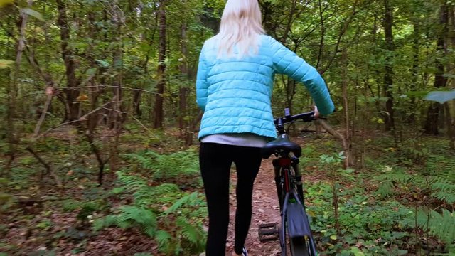 a young athlete in a turquoise jacket and black leggings, slender, pushes a bicycle through the forest in autumn.