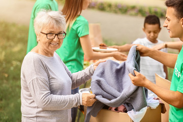 Young volunteer giving new clothes for poor senior woman outdoors