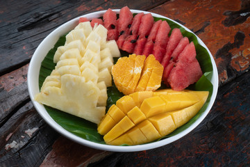 Top view fresh tropical fruits isolated on rustic wooden table. Mango, pineapple, watermelon and orange on a plate with banana leaf
