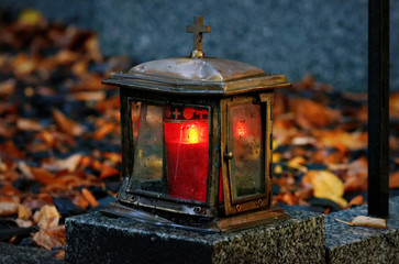 old damaged grave light made of metal with burning candle on a grave with autumn leaves in blurred...