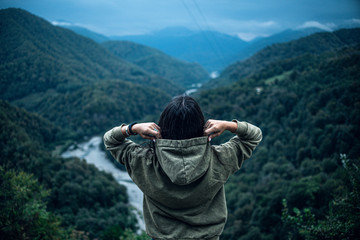 brunette girl in green hoodie puts on hood looks at mountains rear view cloudy sky