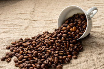 Coffee beans are sprinkled from a coffee cup. Fried arabica beans. Porcelain coffee cup. Linen background. Sackcloth. Nettle fabric.