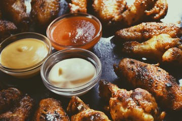 Fried chicken wings. Chicken wings on a dark background on a blackboard. Barbecue meat, grill. Sauce in bowls, ketchup, mayonnaise, mustard, cheese sauce. Appetizing fried meat. Place for your text
