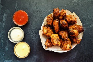 Fried chicken wings. Chicken wings on a dark background. Barbecue meat, grill. Sauce in bowls, ketchup, mayonnaise, mustard, cheese sauce. Appetizing fried meat in the white plate. bowl.