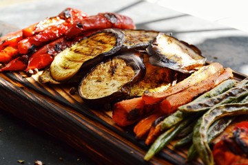 Grilled vegetables on a wooden board. Appetizing grilled vegetables in on a dark background, close-up. Vegetarian wholesome food. Baked eggplant, bell peppers, tomatoes, asparagus and carrots.
