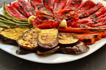 Grilled vegetables on a white plate. Appetizing grilled vegetables in a plate on a dark background, close-up. Vegetarian wholesome food. Baked eggplant, bell peppers, tomatoes, asparagus and carrots.