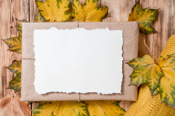 Gift box in kraft packaging. Mockup torn paper for your notes. Autumn bright dry leaves.Yellow knitted sweater on a wooden background. cozy autumn concept. Copy space . flat lay.
