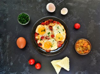 Fried eggs with tomatoes and parsley. Fried eggs in a black plate on a dark background.