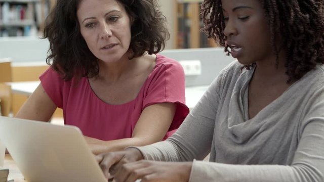 Closeup shot of pensive students working over project at library. Two focused women using laptop while sitting at table. Education concept