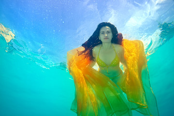 A woman with her hair down swims and poses for the camera underwater in the pool, in a yellow swimsuit, with a yellow cloth in her hands, against the sky. Portrait. Surrealism. Bottom view