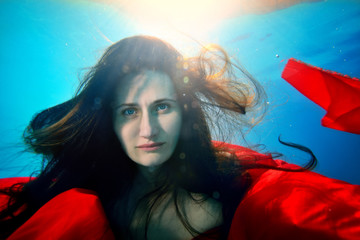 Unusual girl under the water thoughtfully looks at the camera and posing with a red cloth, with her...
