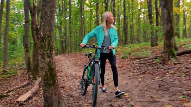 a young athlete in a turquoise jacket and black leggings, slender, pushes a bicycle through the forest in autumn.