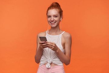 Studio photo of charming young readhead female with bun hairstyle posing over orange background, holding smartphone in raised hands and listening to music with earphones