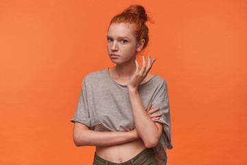 Obraz premium Portrait of attractive young woman with foxy hair in knot standing over orange background, looking to camera confusedly with raised palm. Human emotion facial expression body language