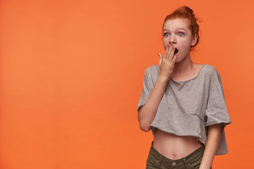 Image of shocked young readhead lady with foxy hair in knot wearing grey t-shirt and green shorts, covering mouth amazedly with raised palm, isolated over orange background