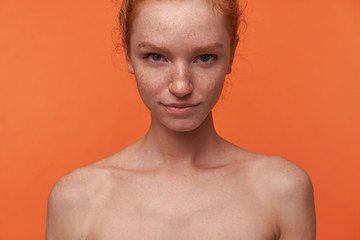 Studio shot of beautiful young lady with red hair in knot standing over orange background with hands down, smiling softly to camera with raised eyebrow. Human emotion facial expression body language