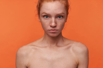 Naklejka premium Indoor shot of pretty young readhead female with casual hairstyle and no make-up standing over orange background, looking at camera with puzzled face, rounding eyes amazedly with folded lips