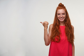 Positive young charming readhead lady in pink dress wearing her red wavy hair in knot, posing over white background with raised thumb and pointing aside, smiling joyfully to camera