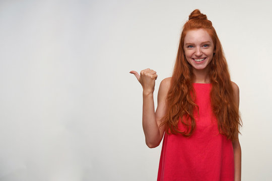 Positive young charming readhead lady in pink dress wearing her red wavy hair in knot, posing over white background with raised thumb and pointing aside, smiling joyfully to camera