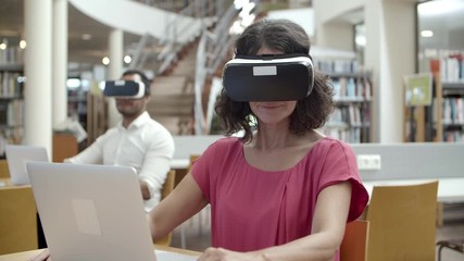 Front view of cheerful Caucasian woman wearing VR headset. Closeup shot of mature user with virtual reality glasses sitting at library. Technology concept - Powered by Adobe