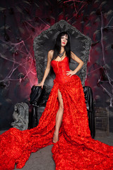 beautiful vampire woman in red long dress near big black throne in the studio