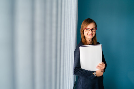 Portrait Of Young Business Woman With Glasses Secretary Holding File Folder Job Application In Front Of Blue Wall By Window Curtains Smiling