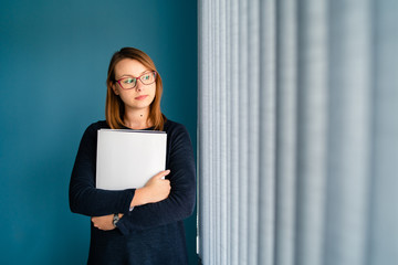 Portrait of young business woman with glasses secretary holding file folder job application in front of blue wall by window looking