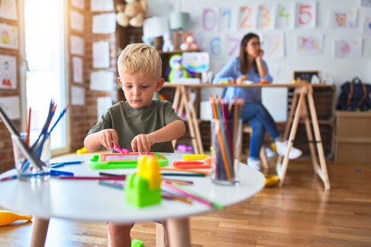 Young caucasian child playing at playschool with teacher. Mother and son at playroom drawing a draw with color pencils, young woman at the background sitting on desk.